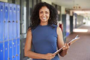 A smiling woman stands confidently with a clipboard, showcasing leadership in front of school lockers.