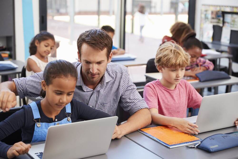 A man and children engaged with laptops in a classroom, highlighting the integration of educational technology in learning.