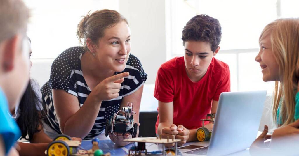 The image shows a classroom scene where a female teacher is interacting with three students around a table. The teacher is pointing something out to the students, who are engaged in a hands-on robotics project. The students, including two boys and one girl, are focused on their work, which includes a laptop and various robotic parts. This setting suggests an educational environment that emphasizes practical, technology-based learning. The teacher’s enthusiastic engagement and the students’ focused expressions highlight an active, collaborative educational process.