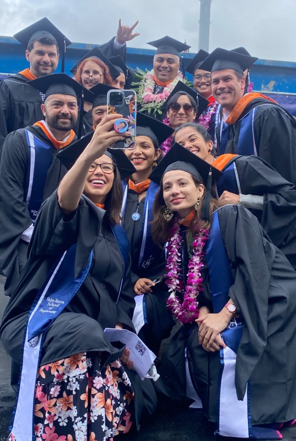 a group of cyber security engineering gradutes in caps and gowns pose together for a selfie after graduation