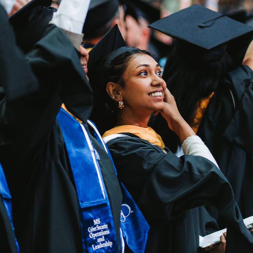 Graduates in black robes and blue sashes celebrate at a commencement ceremony in a large venue.