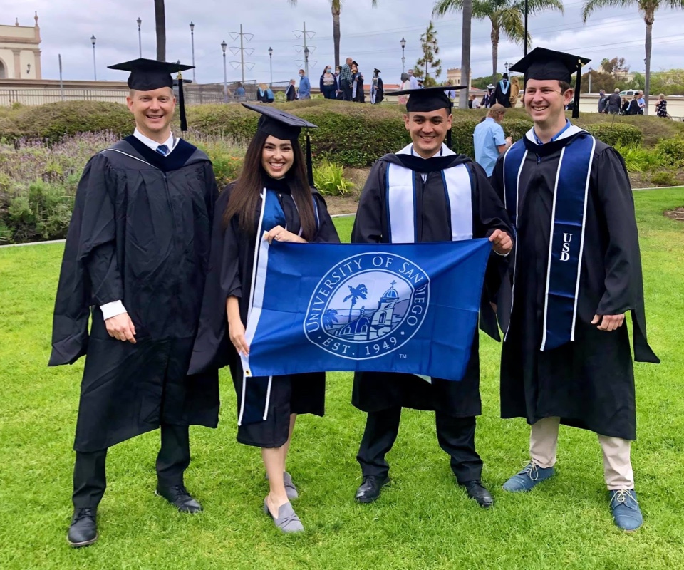 Four USD college graduates in their caps and gowns holding the USD flag in front of them
