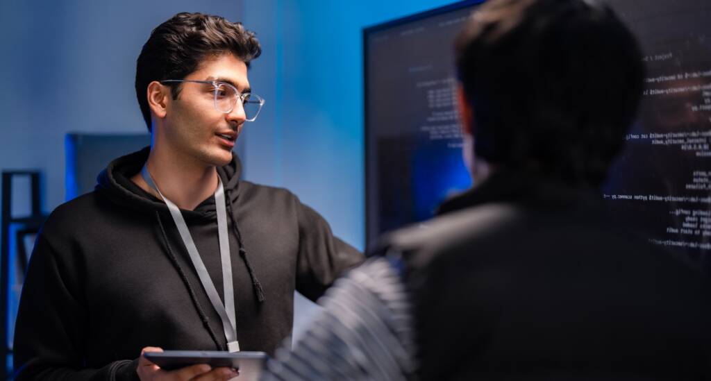 Two Male Tech Software Developers Having a Meeting in a Conference Room Pointing at Computer Screens.
