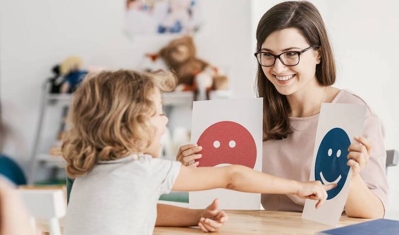 A female teacher is smiling while sitting across a young student holding up an image of a smiley face & a frowny face. The student is pointing to the smiley face.