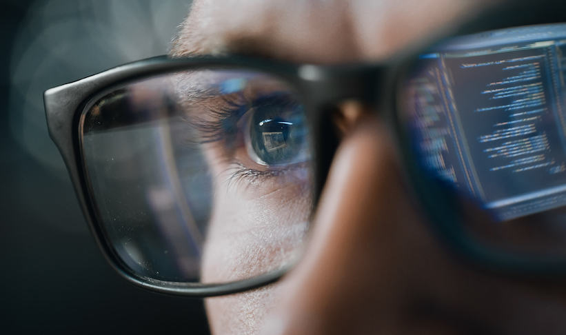 A closeup of man with glasses looking at a computer screen. The screen is reflecting on his left lens