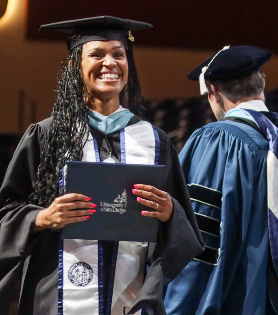 A female USD student in a cap & gown smiles as she holds her new diploma