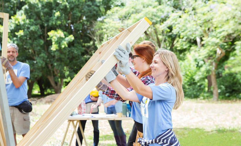 Two women wearing blue volunteers shirts are holding up a wooden frame for a house
