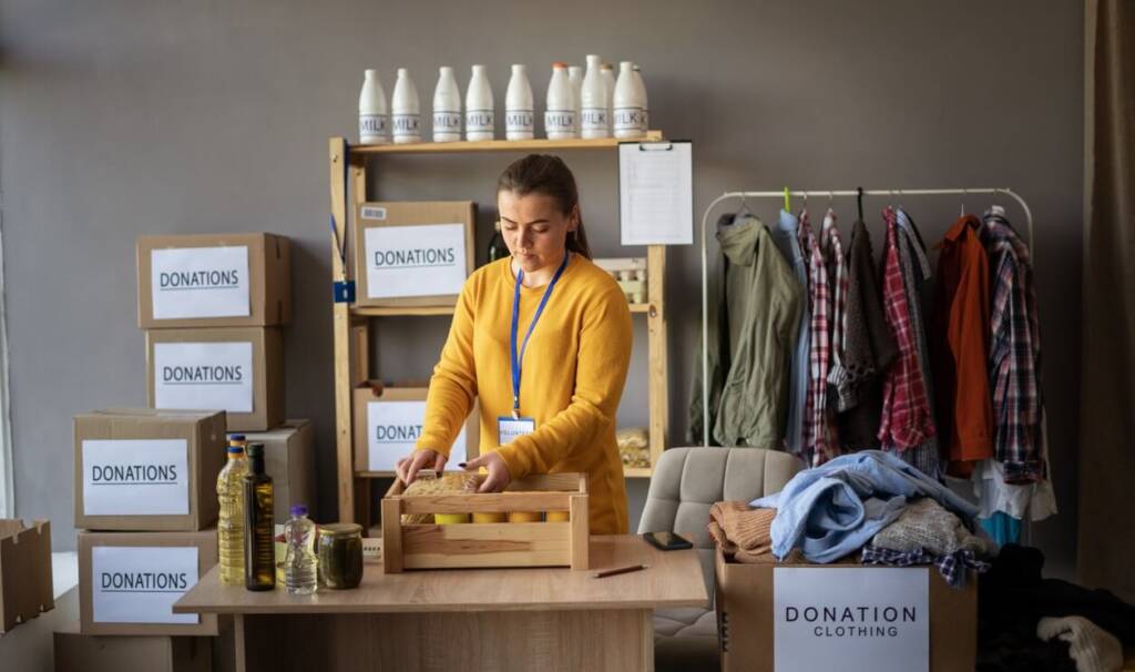 woman with brown hair in a ponytail wearing a yellow sweater and blue lanyard around her neck packs food in a wooden box atop a table, all of which are in front of a wooden rack with boxes labeled "donations" on it