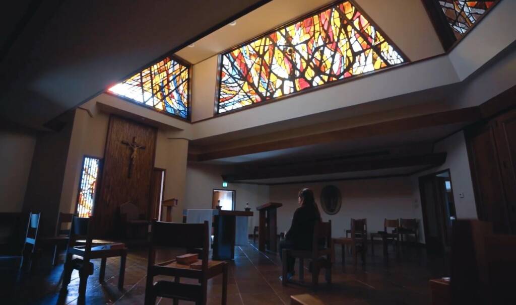 a woman sits in a chair alone in a christian church facing a cross hanging on a wall. there are stained glass windows above the woman and alot of wooden architectural details in the space