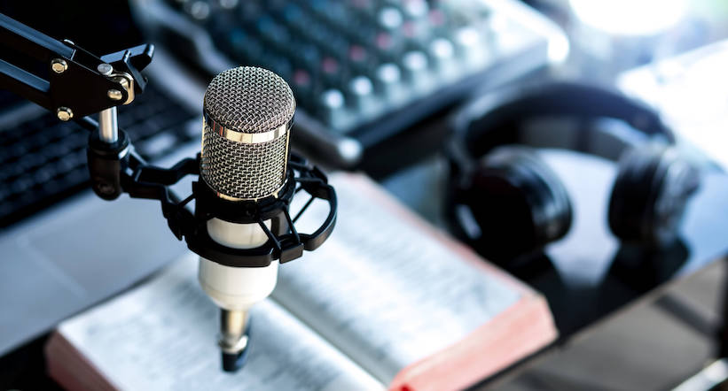 a book lays open on a podcasting desk with a podcasting microphone in the foreground and a mixing board in the background