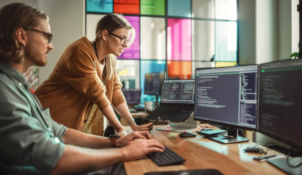 Male AI Trainer and Female Developer Working together at at desk with multiple computer screens with a colorful paneled office window in the background