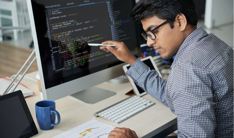 Serious Indian programmer in eyeglasses pointing at computer monitor and checking the statistics of website in document while working at office