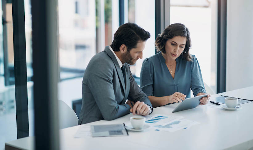 Two business professionals seated at a table, focused on a tablet, engaged in a discussion or collaboration.