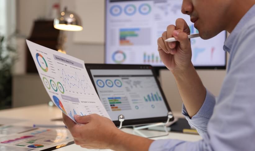 Focused businessman analyzing marketing reports on a desktop monitor and reviewing paper graphs, financial stats, and startup project infographics.