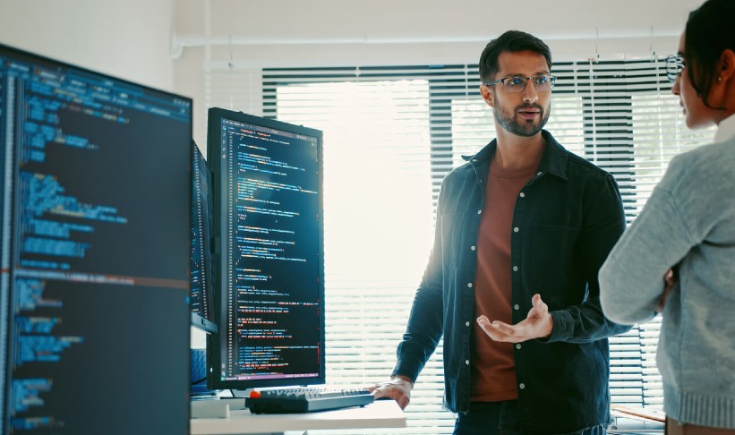 man talking to a woman standing next to monitors with code on the screen