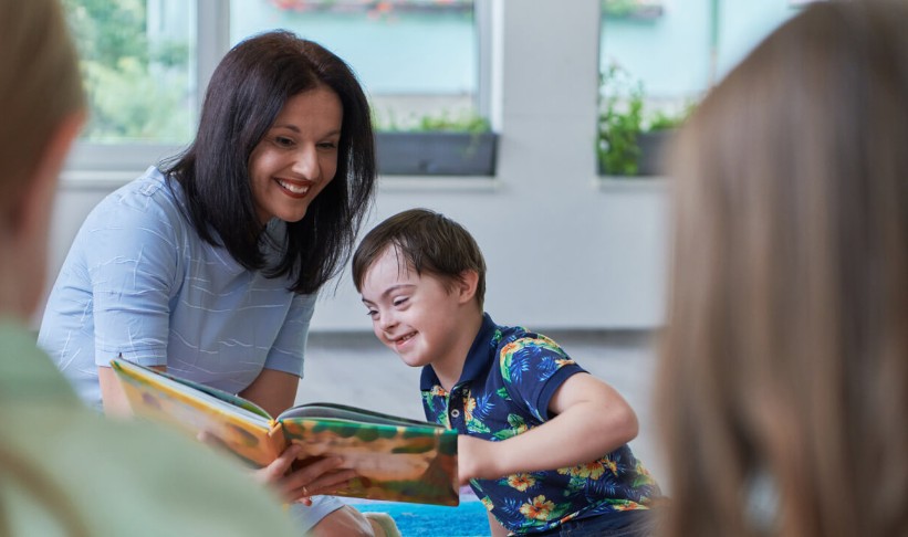 female special education teacher with dark hair and a light blue shirt sits with a male student with dark hair to read a book together