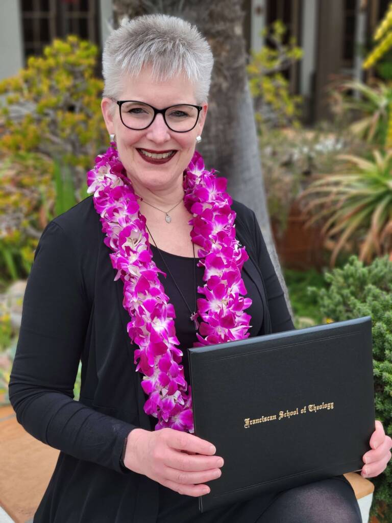 picture of USD PCE alumni Denise Clair, a woman with short grey hair and glasses wearing a pink flower lei and holding her diploma