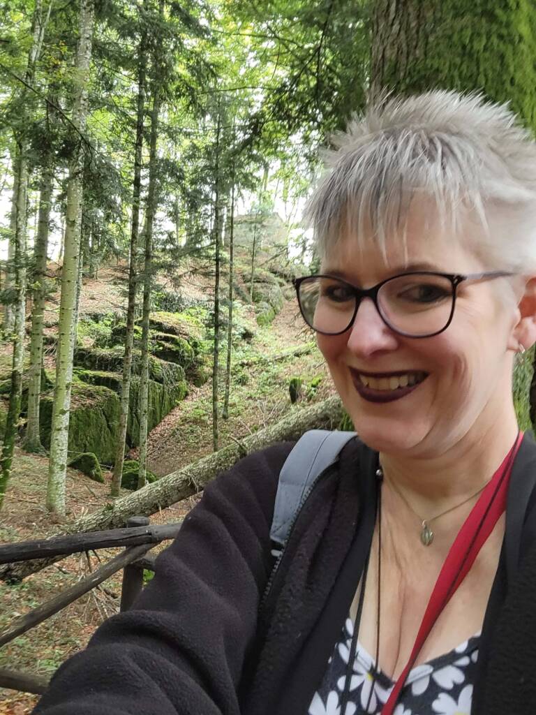 picture of USD PCE alumni Denise Clair, a woman with short grey hair and glasses walking in a wooded area