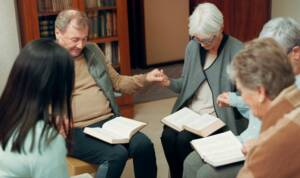 a group of people sit in a circle and hold hands, heads lowered in prayer with bibles on their laps