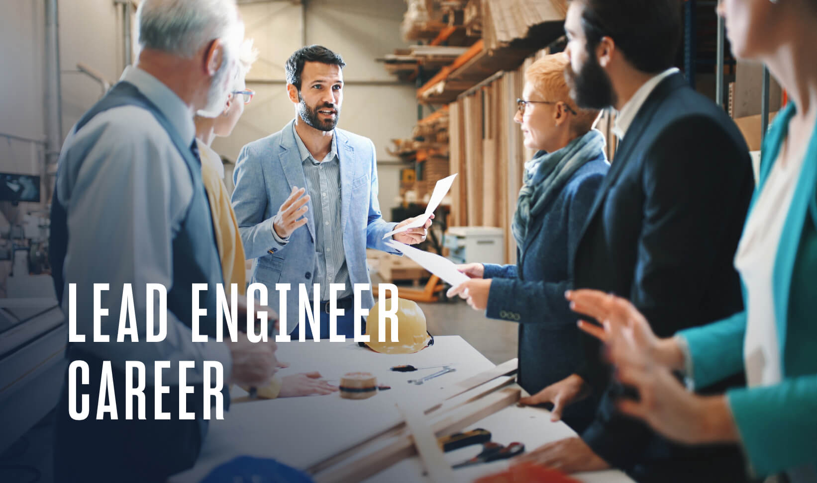 a lead engineer in a grey suit speaks to a group of colleagues at a work desk in a warehouse setting