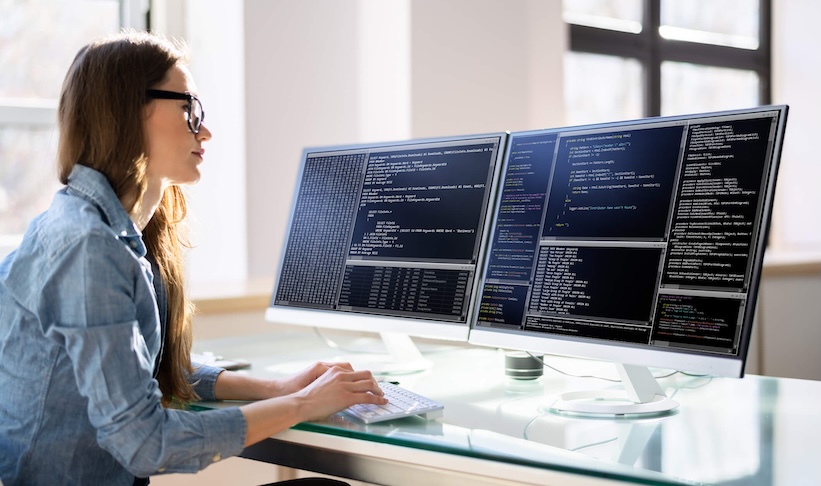 A woman seated at a desk, focused on her work with two computer screens displaying various tasks.