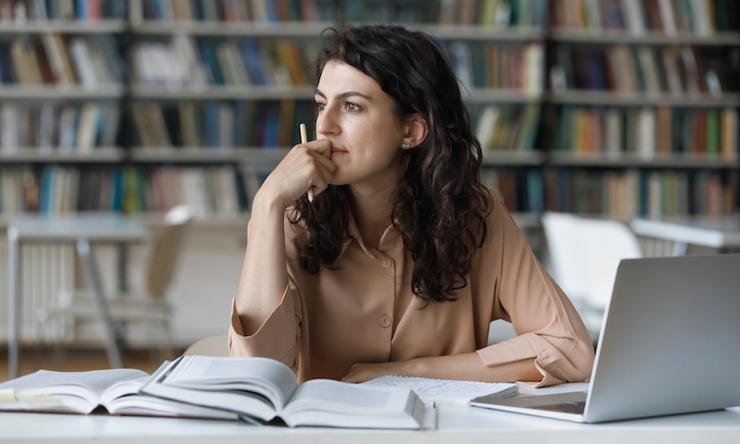 A woman at a table with a laptop open, surrounded by books, deeply immersed in her tasks or research.