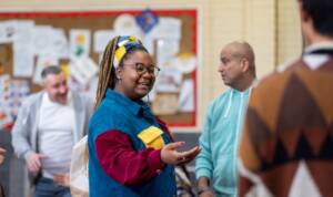 A woman in a vibrant jacket engages in conversation with a group of people, smiling and gesturing animatedly.