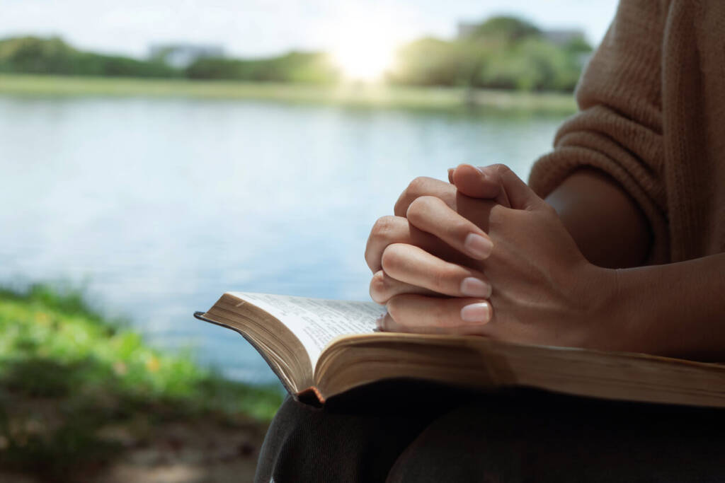 Woman's hands praying with a bible on her legs outdoors