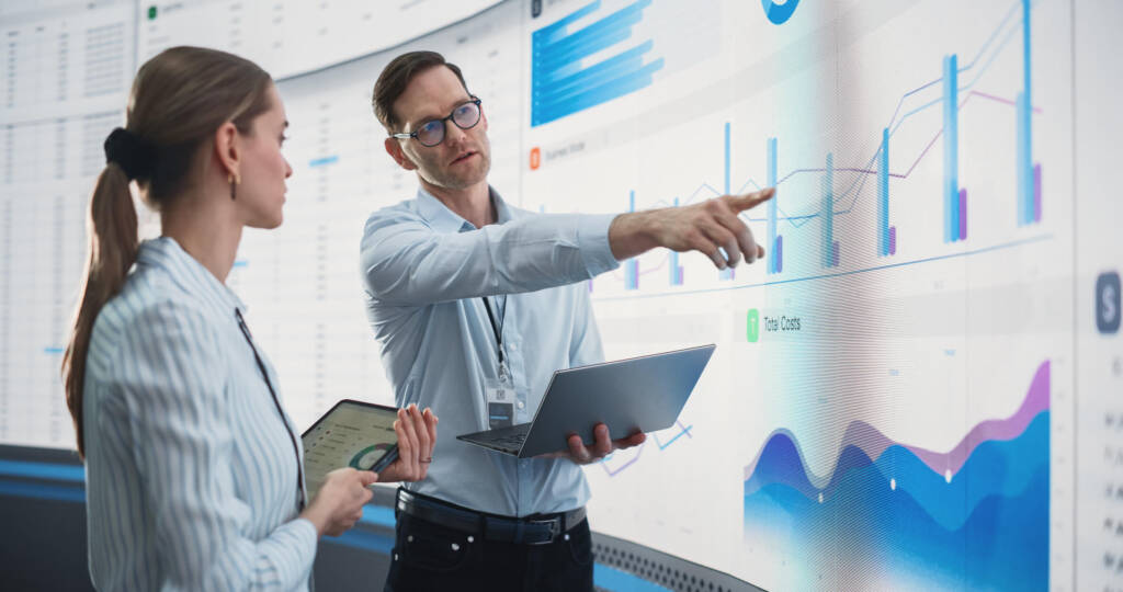 Male And Female Data Scientists With Laptop And Tablet Standing Next To Big Digital Screen With Graphs And Charts