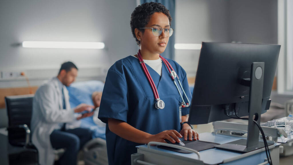 Nurse in scrubs working and looking at a computer in a hospital room