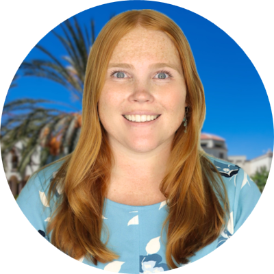 Woman with long reddish-brown hair smiling outdoors with blue sky and palm trees behind her.