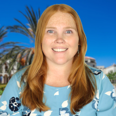 Woman with long reddish-brown hair smiling at camera outdoors with palm trees and blue sky behind.