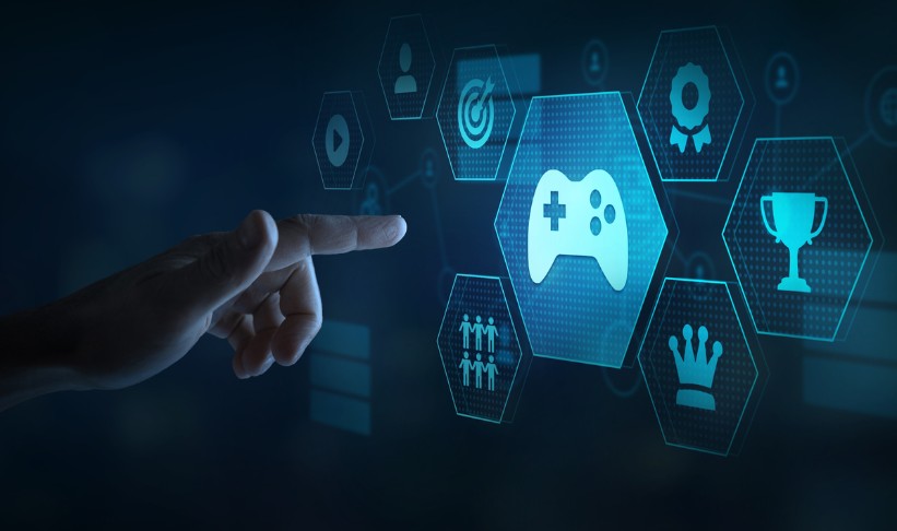 Here's some alt text for the image: Concise version: "A hand reaches toward a glowing blue game controller icon surrounded by hexagonal interface elements representing gamification in learning.