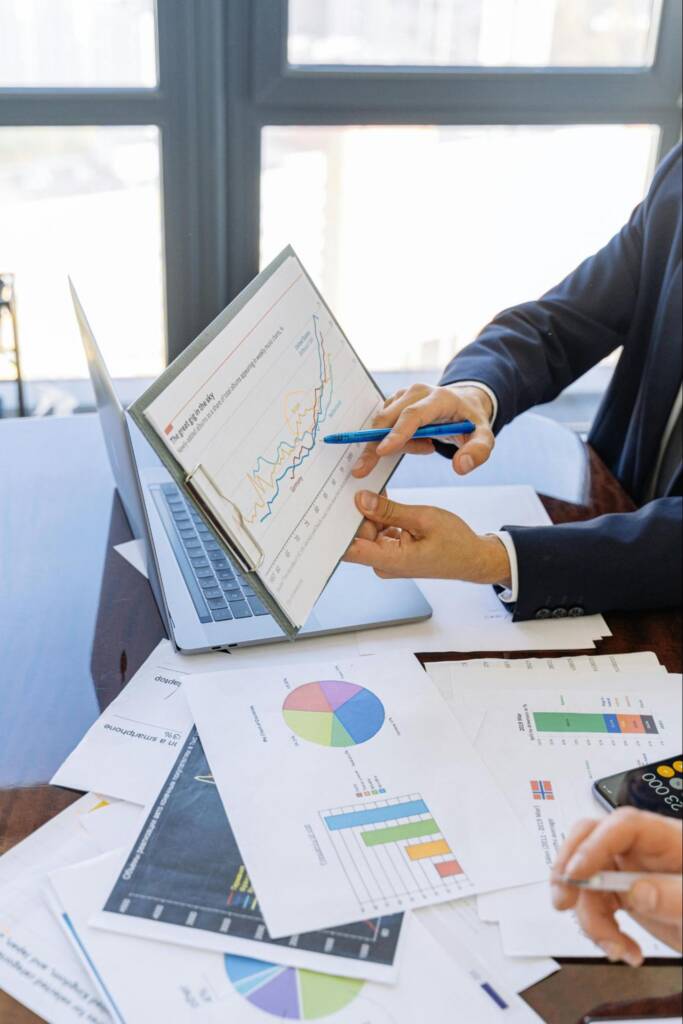 A consultant reviews charts and analytics on a laptop while pointing to a line graph, with printed reports and data visualizations spread across the table.