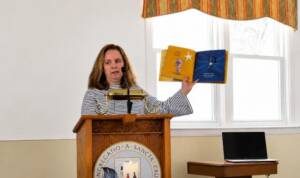 A woman standing at a podium reading a book to a class