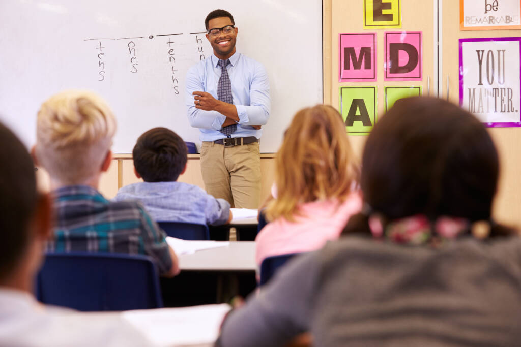 Teacher leaning against a whiteboard, smiling at his students