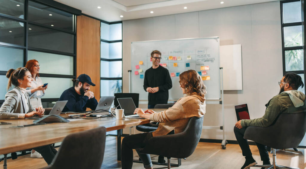 A team of six in a bright conference room smiling during a meeting, while one man stands at a whiteboard covered in sticky notes.